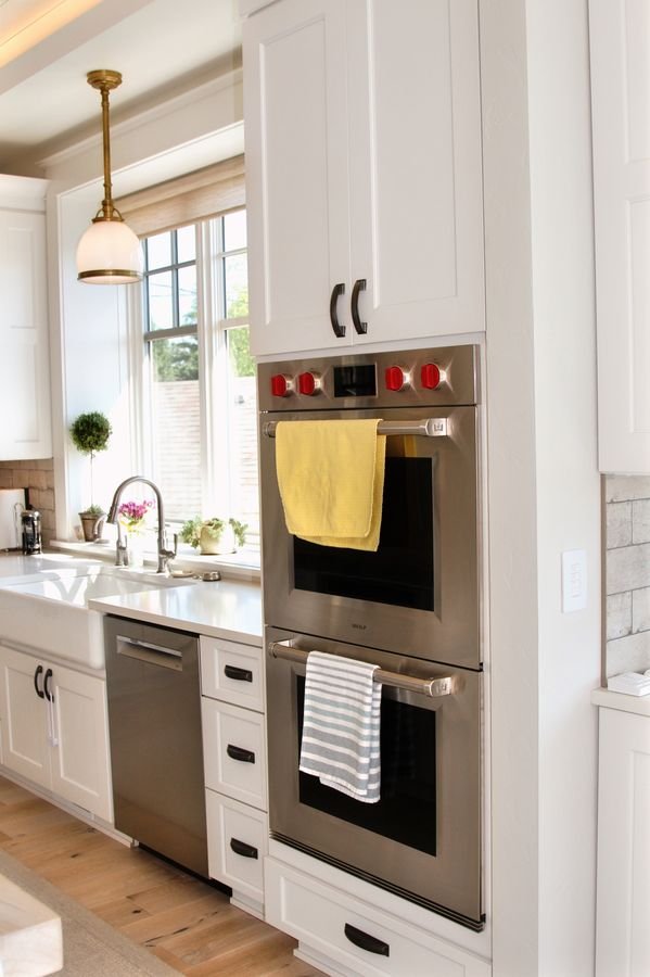 Stainless double wall ovens set into white cabinetry, with the sink and a pendant light beyond