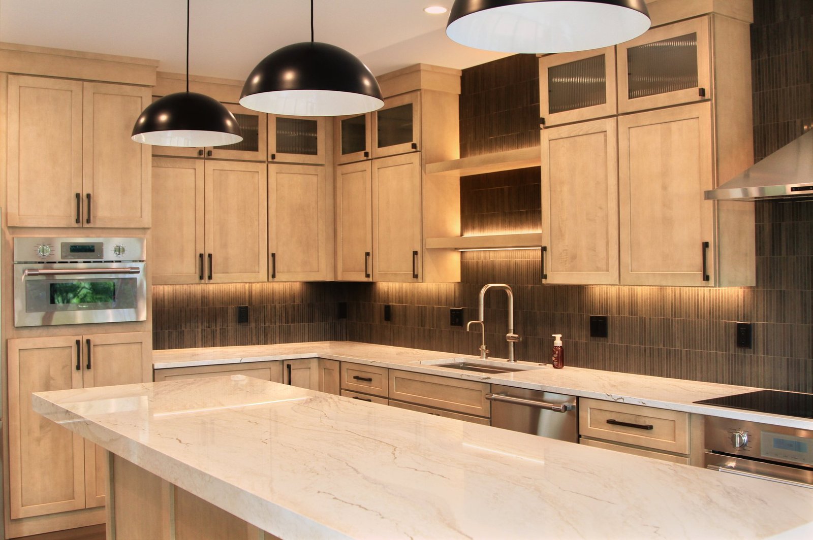The Whitewater Lakehouse kitchen: long stone island under black pendants, maple cabinetry, and a dark tile backsplash