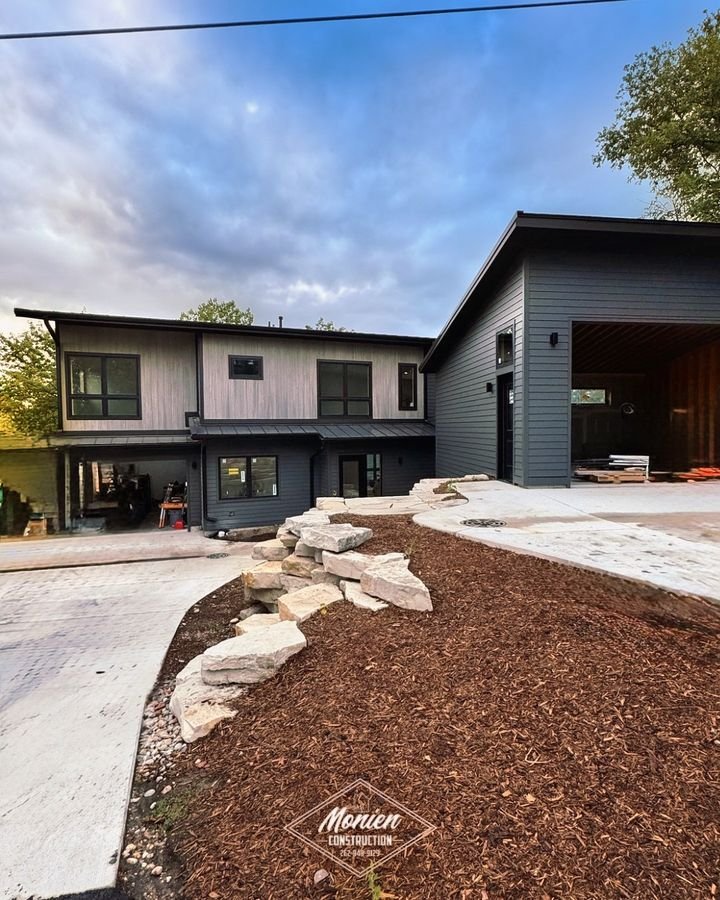 Front elevation with light wood siding above dark siding and a natural stone outcrop beside the drive